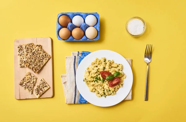 Tasty scrambled egg with tomato and basil in white plate over kitchen towel. Fork, sea salt, Crispbread and fresh raw chicken eggs in egg carton box on yellow background. top view