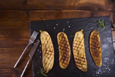 Çatal ve bıçak koyu kahverengi rustik ahşap arka plan üzerinde siyah arduvaz plaka üzerinde sırlı patlıcan ızgara. Üstten Görünüm