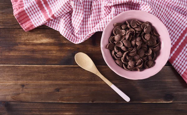 Chocolate cereals in pink bowl, spoon and kitchen towel over dark brown wooden background. Top view