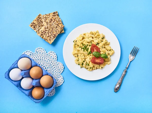 Tasty scrambled egg with tomato and basil in white plate. Crispbread and fresh raw chicken eggs in egg carton box over beautiful napkin on blue background. top view