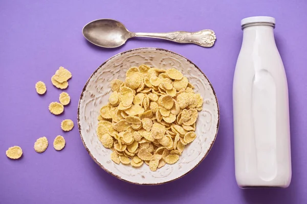 Cereals in bowl, spoon and bottle with milk over purple background. Top view