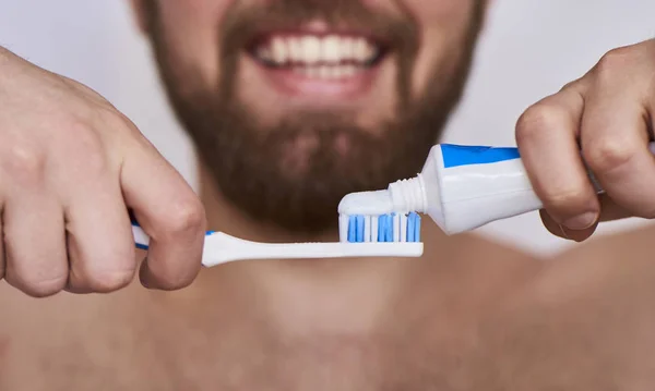 歯ブラシを持つ男 歯磨き粉と歯ブラシを押しながらニコニコと灰色の背景に対して立っている若い男の画像をトリミング — ストック写真