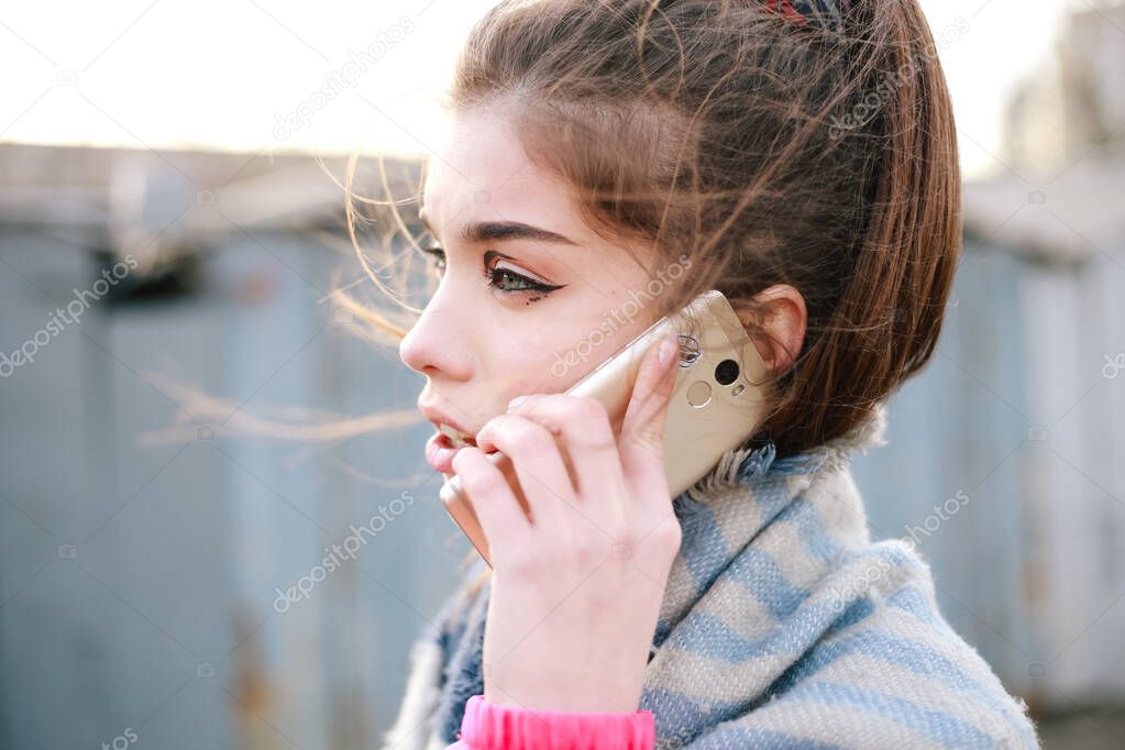Candid portrait of young woman \ girl in warm scarf talking on phone worried, concerned and emotional looking away near garages. Street style, wind blows hair