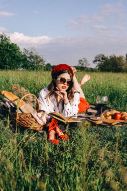 Doğada modern yaz pikniği. Kırmızı şapkalı güzel bir kız ve güneşli bir günde siyah güneş gözlüklü. Kitap okuyor ve anın tadını çıkarıyor.