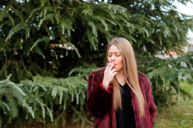 sarışın genç kadın parkta sigara kırmızı kürk palto giyiyor