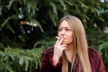 sarışın genç kadın parkta sigara kırmızı kürk palto giyiyor