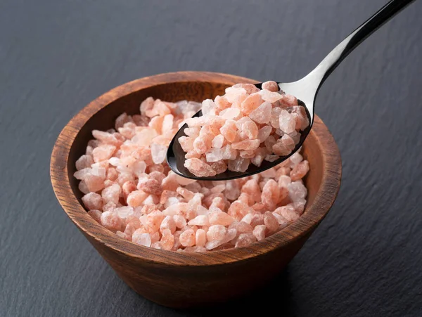 Lifting pink rock salt in a wooden bowl on a black background with a black spoon