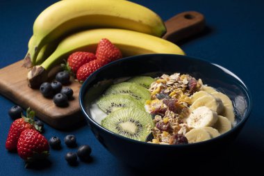 Meyve, granola ve yoğurt koyu mavi arka planda bir kabın içinde servis edilir. Yanına bir muz, çilek ve yaban mersini..