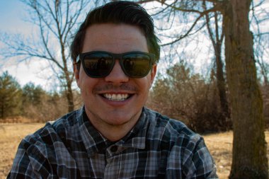 Gülümseyen genç bir adam, modaya uygun, ekose bir gömlek giyiyor ve arkasında yeşil bir kör var. Sakallı ve şehir arka planında modern saç stili olan adam.