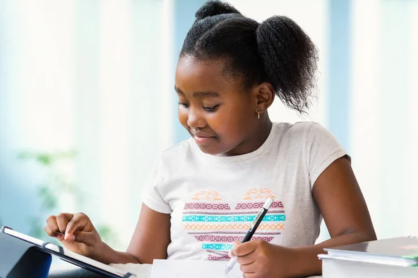 关闭可爱的小非洲女孩的肖像做家庭工作在办公桌上 Ponytailed 儿童在数字平板电脑上打字和用钢笔写字 — 图库照片