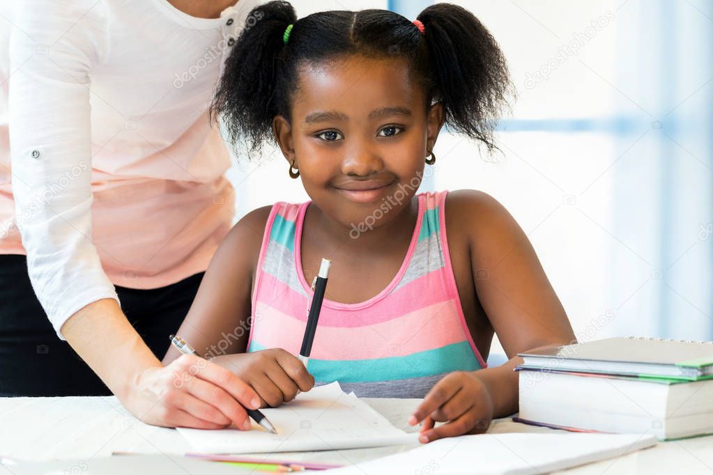 Close up portrait of cute little african student doing homework with caucasian teacher. Ponytailed girl sitting at desk with pen,paper and books.