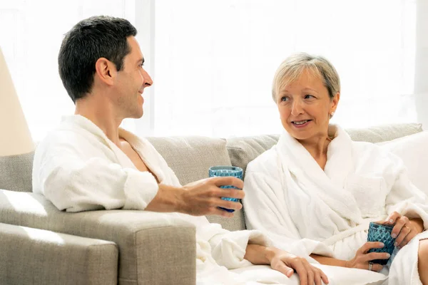 近照中年夫妇在家里聊天的肖像 穿着白色浴衣的情侣们坐在沙发上举着眼镜 — 图库照片