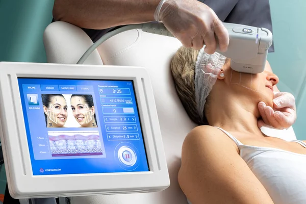 Middle aged woman having skin tightening ultrasound treatment on cheek. Digital display on hifu equipment with therapist and patient in background.