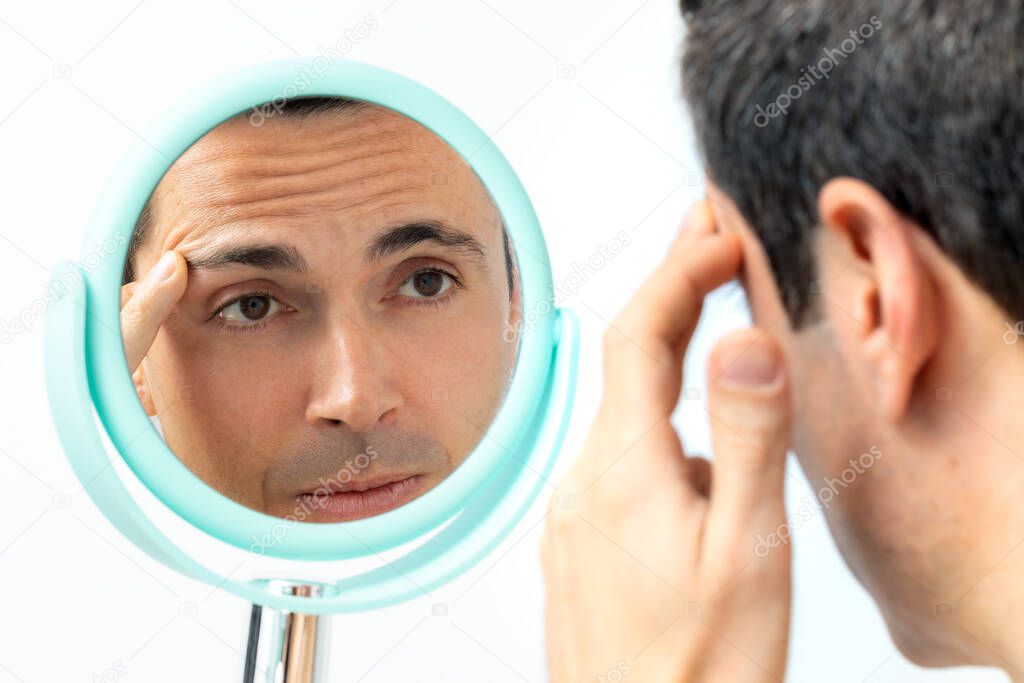 Close up portrait of middle aged man reviewing wrinkles and sagging skin in hand mirror. Isolated on light background.