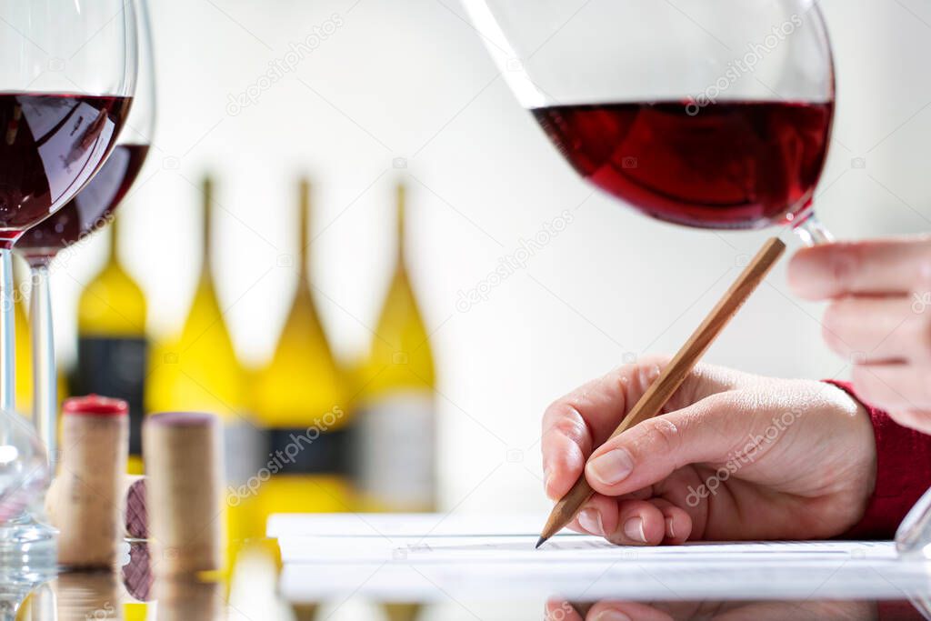 Close up detail of enologist writing notes on wine evaluation at table. Out of focus bottles in background.