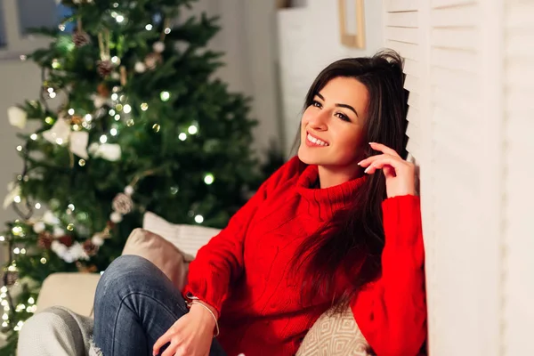 Una chica sonriente cómodamente sentada en un sofá entre almohadas cerca de t —  Fotos de Stock