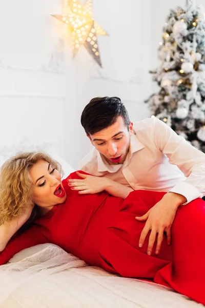 young parents felt the jolts of their future baby lying on a bed on the background of a Christmas tree