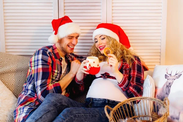 Les futurs parents dans le chapeau du Père Noël sur un canapé manger des biscuits — Photo