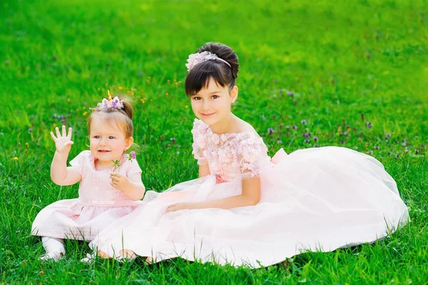 Little sisters are sitting on a grass in a park in festive dress — Stock Photo, Image