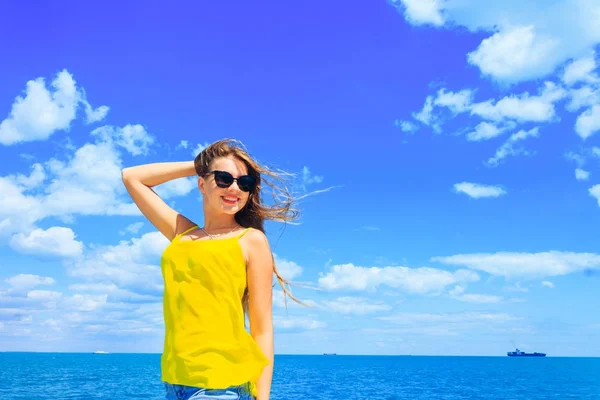 美丽的年轻女孩穿着黄色上衣和太阳镜对海 船和蓝天的背景微笑 — 图库照片