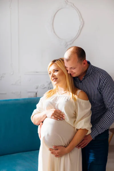 Muž Objímá Svou Těhotnou Manželku Pozadí Pohovku Zdobené Bílou Zeď — Stock fotografie