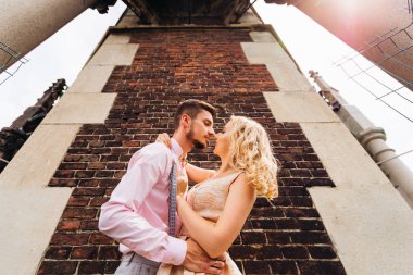 Çocuk kız arkadaşının beline sarılıyor ve onu tuğla duvarın yanında öpmek istiyor.