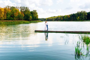 Birbirine aşık bir çiftin, sonbahar ağaçlarının kıyısındaki pitoresk bir gölün iskelesinde nazikçe kucaklaşmaları.
