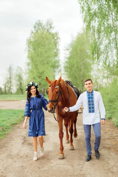 一个穿着礼服 头上戴着花圈的女孩和她的男朋友 穿着一件绣花衬衫 走在土路上 拿着一匹马当马 — 图库照片