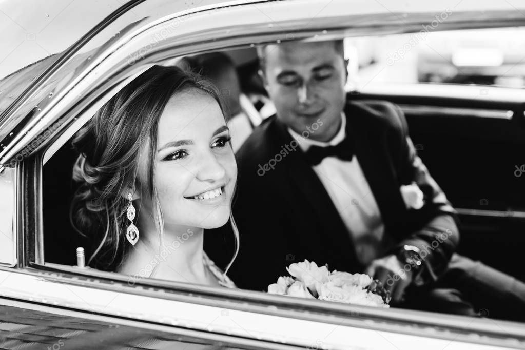 newlyweds are sitting in a black car, the bride smiles and looks through the window on black and white photography