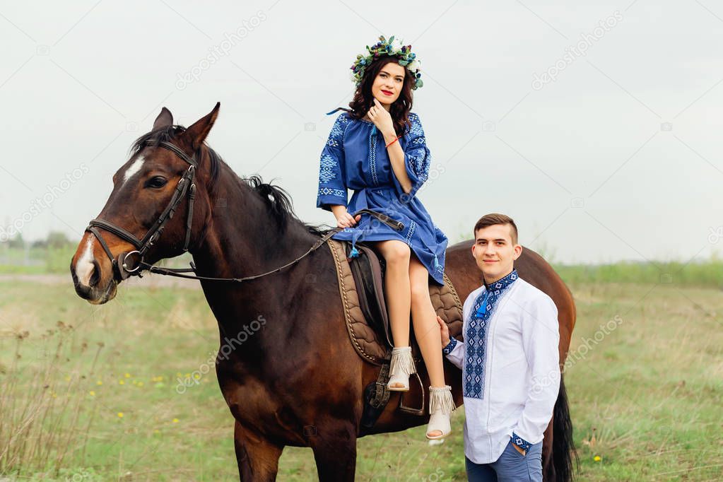 The girl elegantly sits on a horse and her boyfriend stands next to her and they look together in the camera lens