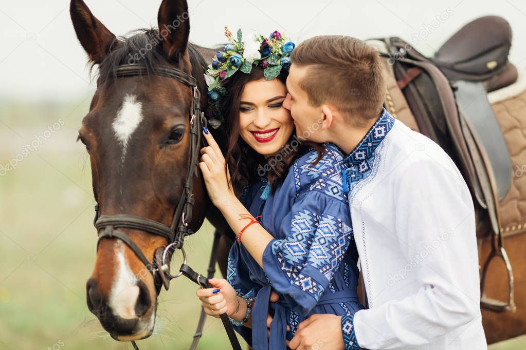The guy kisses his girlfriend with a wreath on her head that closes her eyes and smiles and holds the horse for a bridle