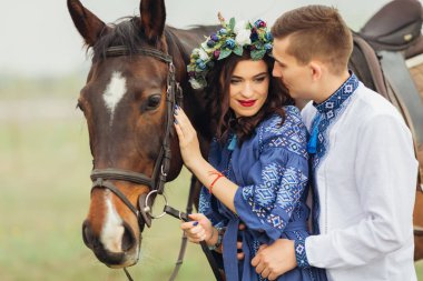 Kafasında güzel bir çelenk olan hamile bir kız bir atı dizginler ve onu kucaklayan sevgilisinin yanında durur.