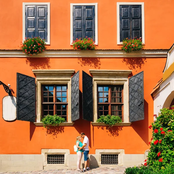 A magnificent building with an orange facade and large windows on the windowsill of which are beautiful flowers. A mature couple stands face to face near this building