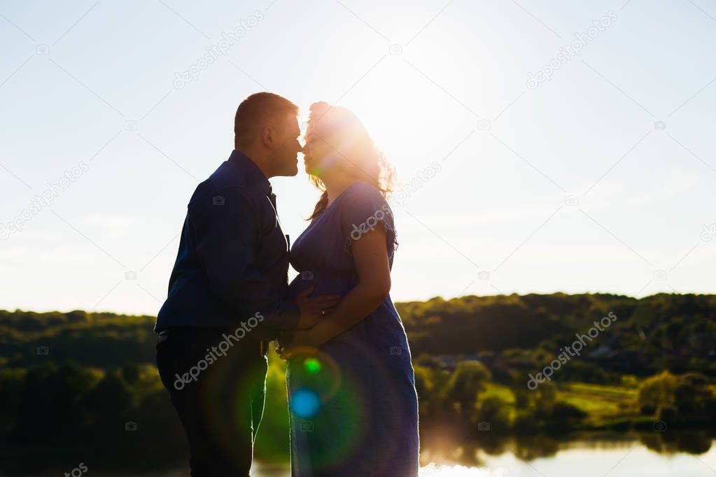 A man kisses his pregnant wife on the background of a small lake