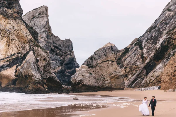 large rocks by the ocean and sandy shore. the newlyweds are hold