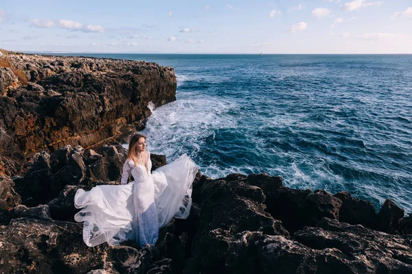 Noiva está entre rochas com vista para o oceano. vestido voando. — Fotografia de Stock
