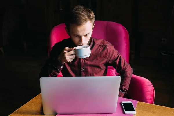 the guy drinks tea and sits in armchair. computer and phone on t