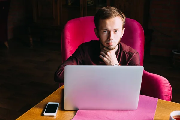 bearded guy looks into the camera and sits on armchair. laptop a
