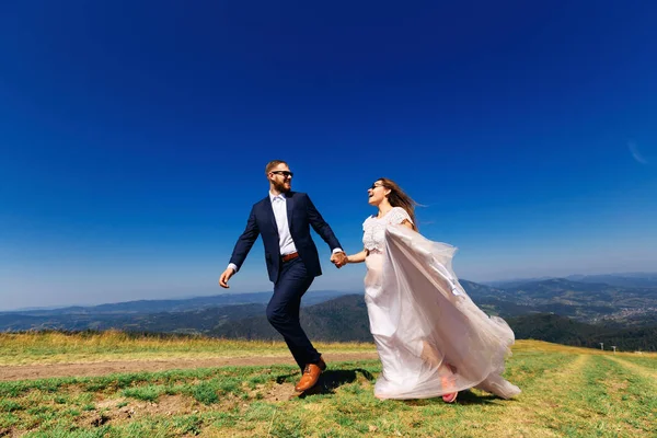 喜庆的新婚夫妇在山顶的草丛中奔跑着。 — 图库照片