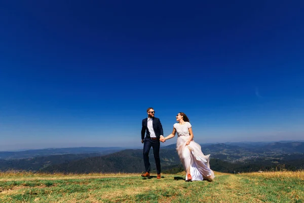 Recém-casados de mãos dadas e correr em grama verde no topo da montanha — Fotografia de Stock