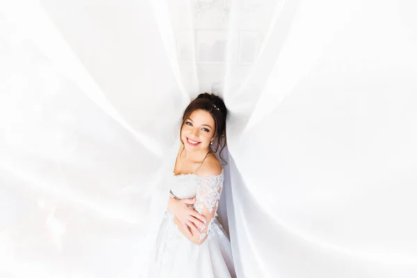 Невеста в белом платье улыбается и обнимается под белым занавесом — стоковое фото