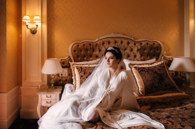bride in white dressing gown and bridal veil sitting on chic bed