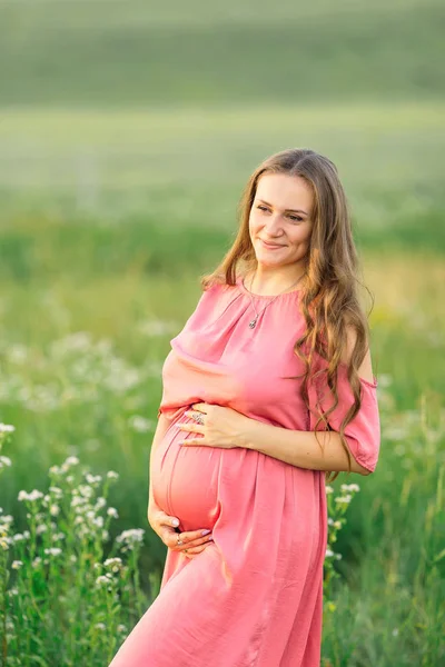 Zwanger vrouw in roze jurk raakt buik en kijkt in de camera — Stockfoto