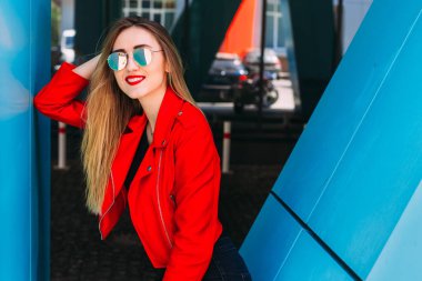 Deri ceketli ve güneş gözlüklü genç bir kızın portresi şehrin modern binasının yakınlarında poz veriyor. Kapat..
