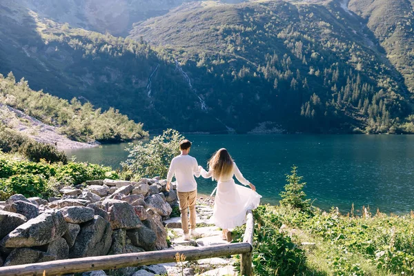 女孩和男人牵着手走在石路上 湖与山的美丽景色 后视镜 — 图库照片