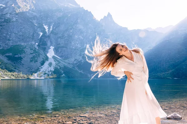 湖の近くの白いドレスの少女は手を腰に当てて髪を振っている 山と太陽 — ストック写真