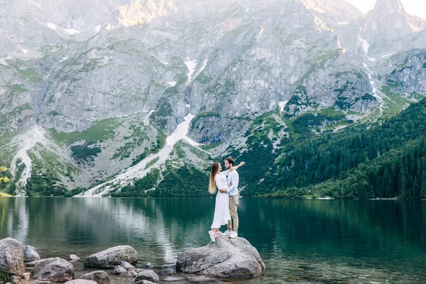 情侣们拥抱在一起 站在湖水和高山的背景下的石头上 — 图库照片