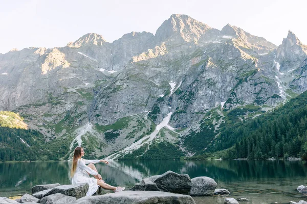一个身穿白衣的女孩的肖像坐在一块大石头上 欣赏高山和湖景 — 图库照片