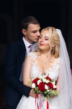 Zarif elbiseli güzel gelin düğün buketi taşıyor. Şık takım elbiseli damat geline arkadan sarılıyor..