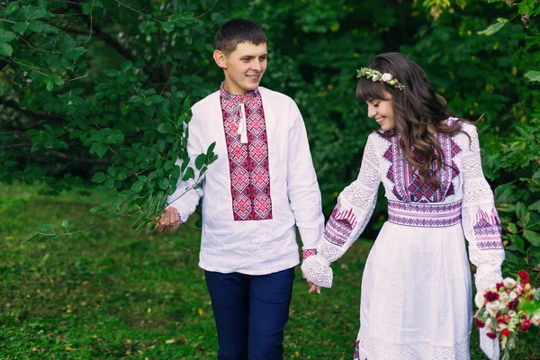 刺繍入りの服を着た新婚夫婦が手を取り公園を歩く ウェディング花束を持った花嫁 — ストック写真